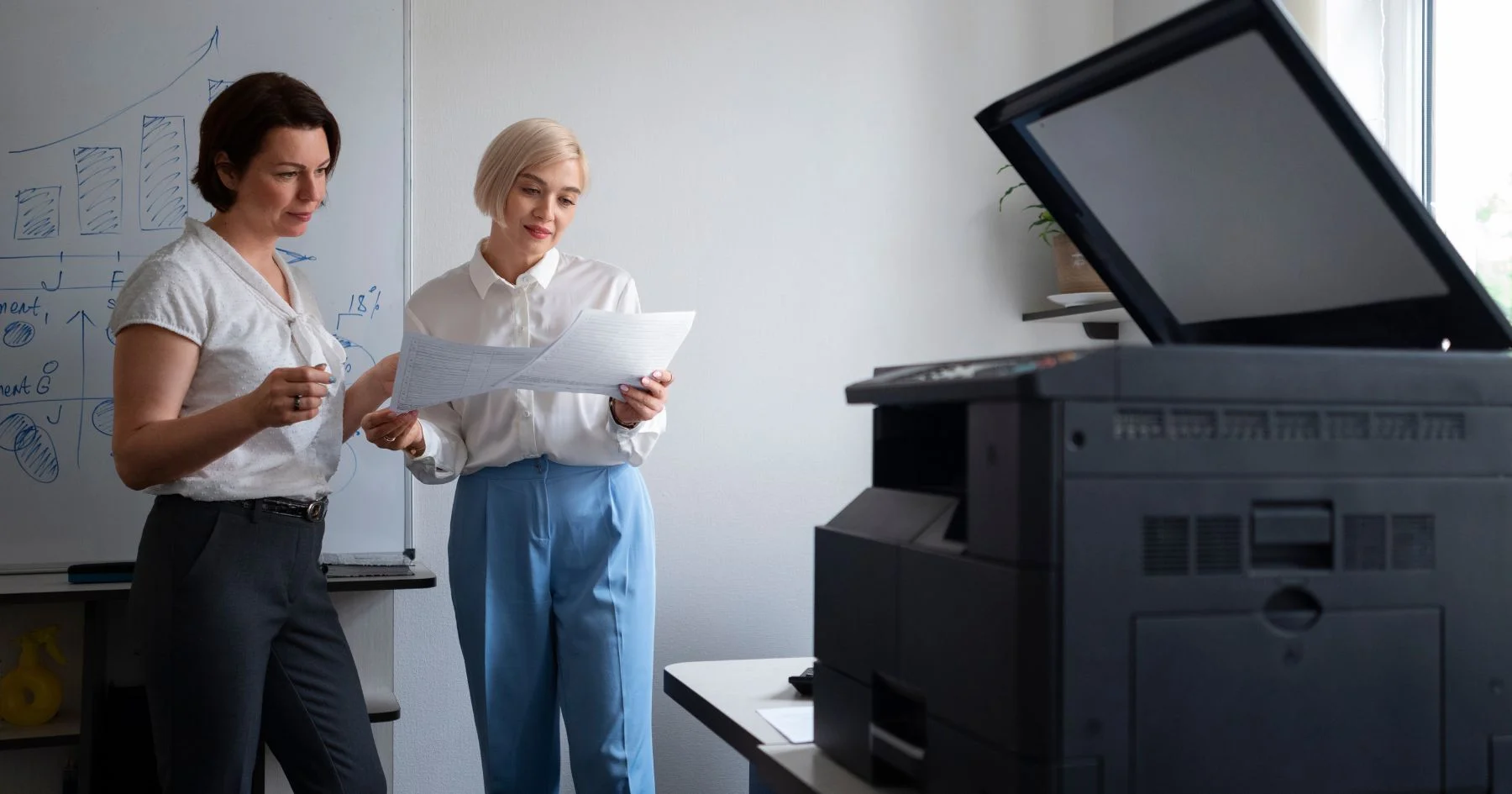 Two businesswomen reviewing documents beside office printer