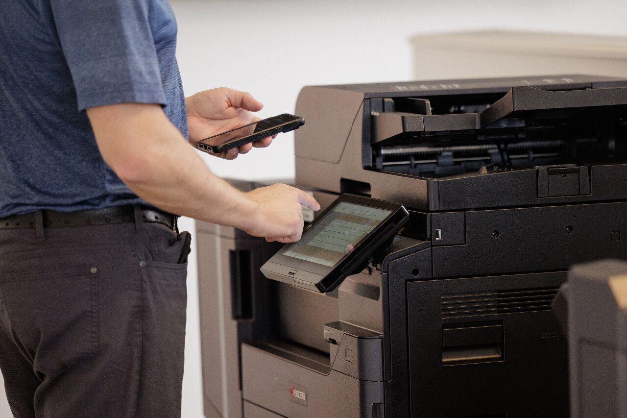 a man standing next to a kyocera multifunction printing with his mobile device in his hand